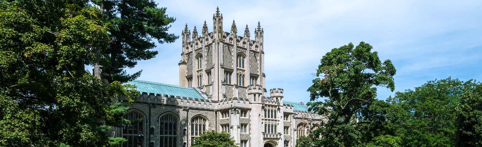 A gothic stone building with tall spires visible above a treeline on a clear, sunny day.